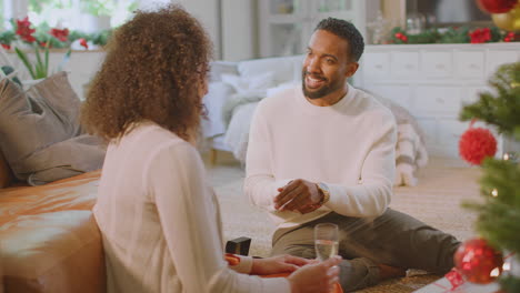 loving couple drinking champagne as man tries on gift of wristwatch around christmas tree at home