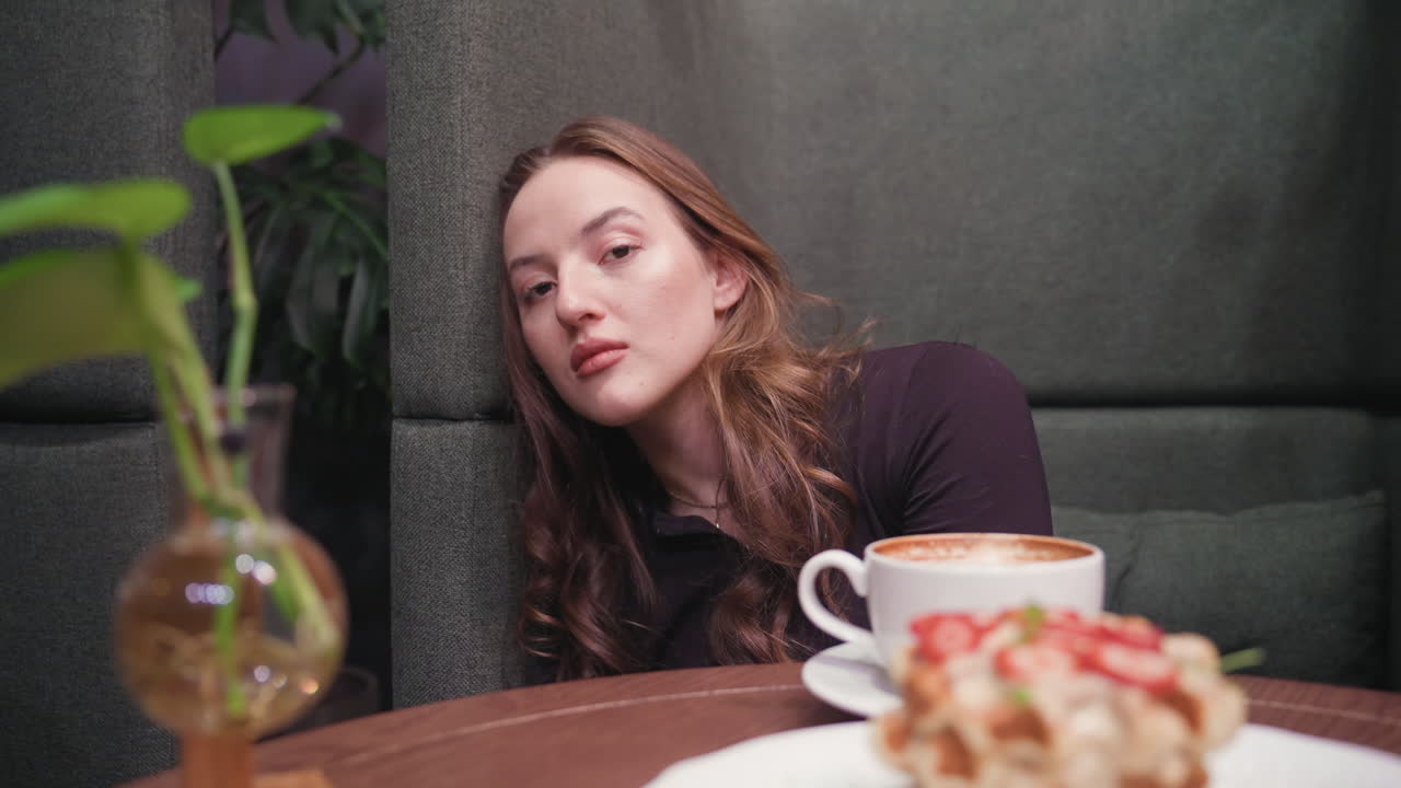 Young woman with long wavy hair leans head on couch wall looking calm and relaxed in cozy cafe, table holds white tea cup, dessert with strawberries, and decorative glass vase with green plant
