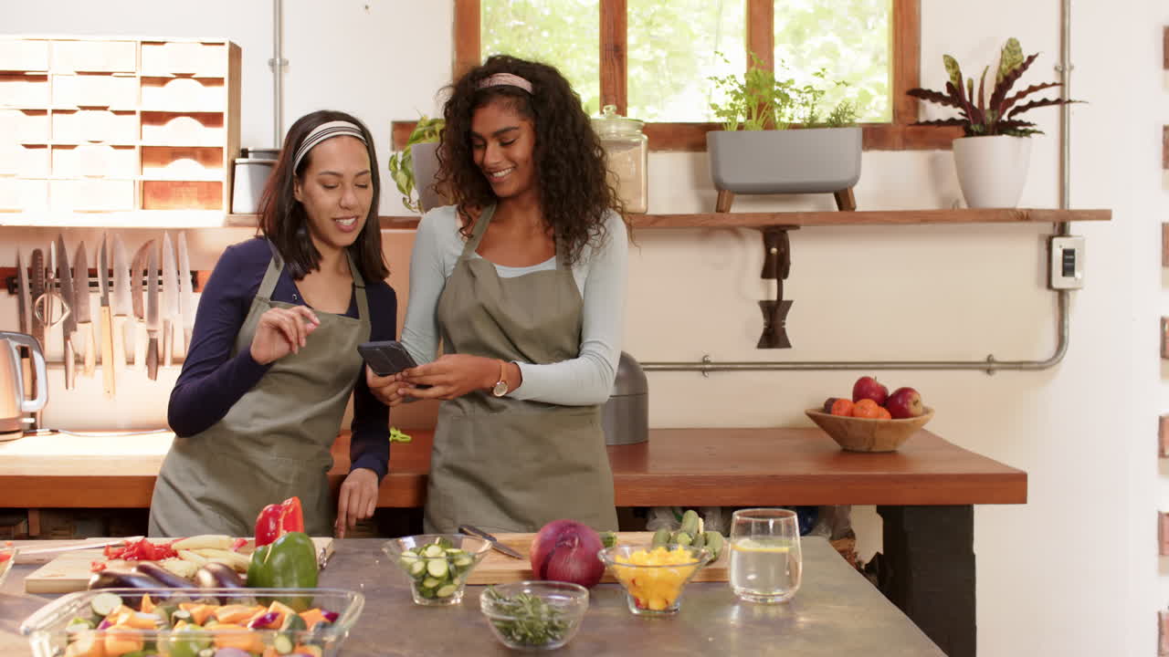 Two multiracial female friends in kitchen sharing smartphone while preparing vegetables, at home