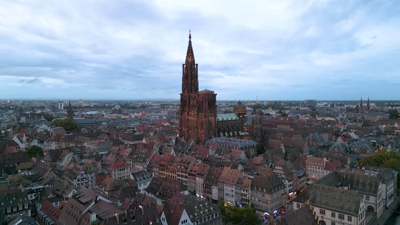 Veduta aerea al crepuscolo di Strasburgo con la Cattedrale di Strasburgo illuminata che si erge sopra gli incantevoli tetti del centro storico, sotto un cielo blu sereno.
