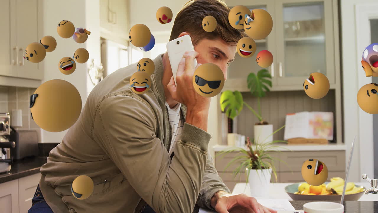 Man leaning in modern kitchen, holding smartphone while checking laptop with floating tech emojis