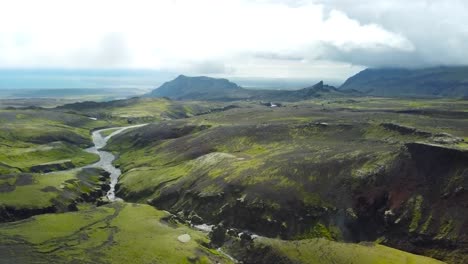 aerial drone footage flying over green mossy ruged and dark rocky volcanic dark brown terrain landscape mountains in iceland with a small narrow river flowing in the middle of the land. sun shining.