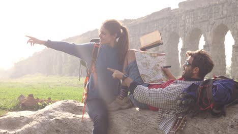 pareja feliz de mochileros turistas sentados acostados en un tronco de madera leyendo una guía de mapas apuntando a las antiguas ruinas del acueducto romano en el parco degli aquedotti parque en roma al amanecer romántico con saco de dormir cámara lenta