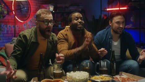 three mixed-races male friends resting in the evening at home with beer and cheering up for a goal on the couch in front of tv with sport channel.