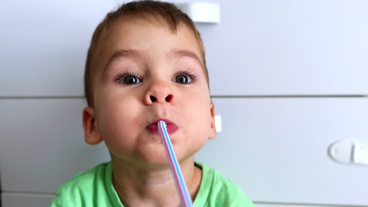 Adorable face of a Caucasian toddler holding a plastic straw in his mouth. Baby boy drinking from a straw. Close up. Vertical screen.