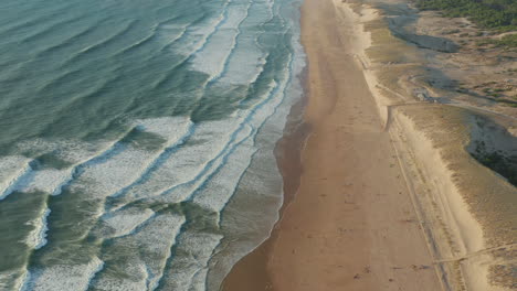 Few-People-on-wide-Brown-Beach-keeping-distance-at-Beautiful-Golden-Hour-with-White-Green-Waves-Crashing-on-Shoreline,-Aerial-Wide-View-slow-tilt-up