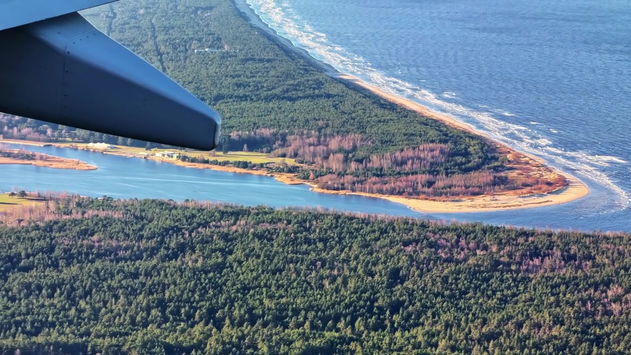 Lentokoneen ikkunakuva näkyvällä siivellä Itämeren rannikon yllä, paljastaen hiekkaisen niemen, tiheän metsän, aallot ja sinisen meren lähellä Riikaa, Latviaa.
