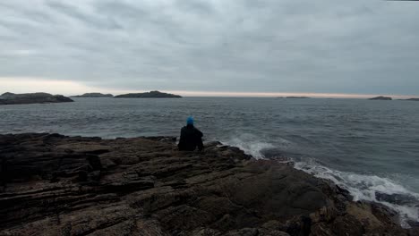 man sitting on cliff close to ocean in moody light after sunset with waves crushing up on shore around him - static clip with thoughtful person and dramatic cloudy sky