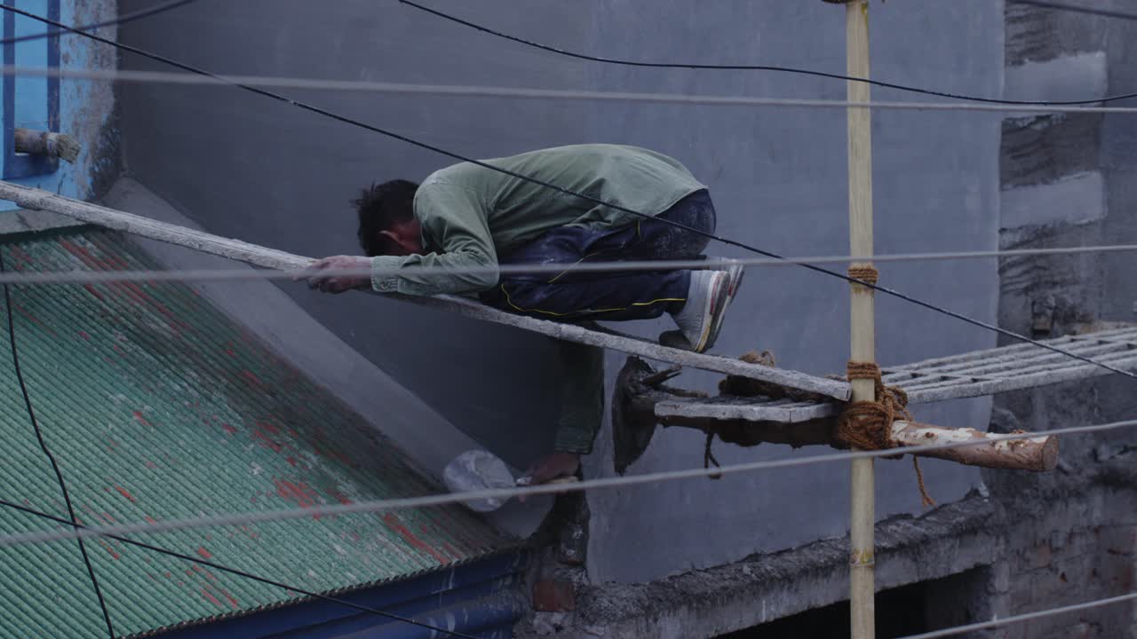 A slow motion handheld long wide shot of an Indian man wearing faded olive green shirt, working at a construction site, applying cement on roof while sitting on a thin pole at a height