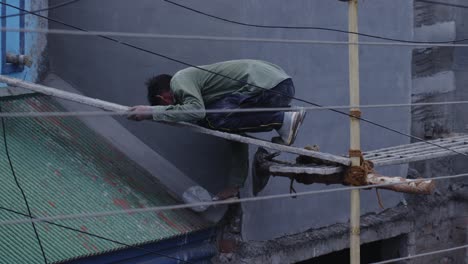 una foto larga y lenta de un hombre indio con una camisa verde oliva desvanecida, trabajando en un sitio de construcción, aplicando cemento en el techo mientras estaba sentado en un poste delgado a una altura