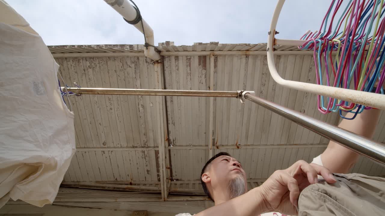 한 아시아 남자는 세탁물과 젖은 옷을 차양 아래에서 말리기 위해 걸어둔다 - 느린 동작으로 위를 바라보는 낮은 각도
