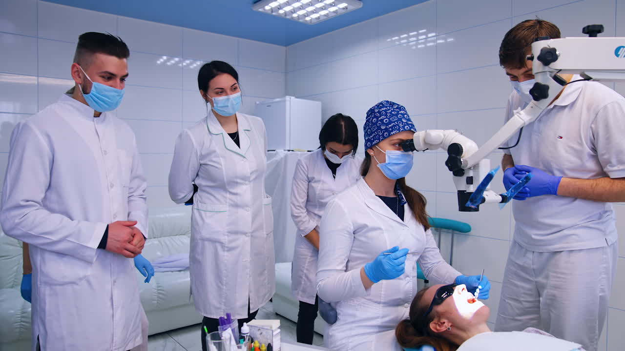 Brunette student in cap and mask sits in front of microscope. Teacher stands beside and students are at backdrop.