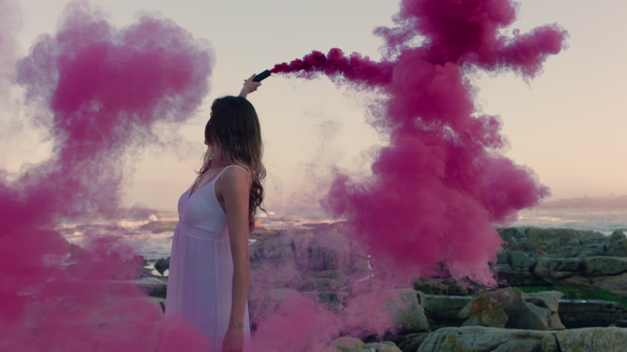 bellissima donna che agita una bomba di fumo rosa che balla sulla spiaggia all'alba celebrando la libertà creativa