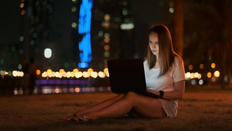 A-woman-software-developer-sits-in-a-Park-on-a-summer-night-in-the-city-and-writes-code-looking-at-a-laptop-screen.-Print-the-article-on-a-laptop.-Blogger-replies-to-posts