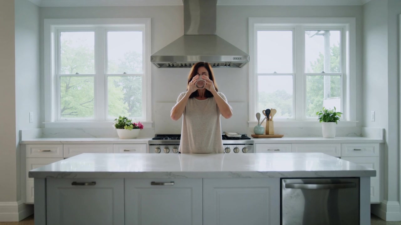 Donna alza una tazza, sorseggia e l'abbassa all'isola della cucina, illuminata dalla luce del mattino proveniente dalle finestre.