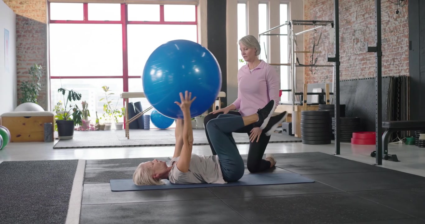 Ein Trainer gibt einem älteren Klienten Anweisungen, der im Studio Bein- und Armübungen mit einem blauen Ball zur Stärkung der Rumpfmuskulatur ausführt.