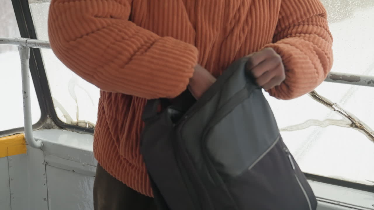 Uomo di colore sul tram che apre lo zaino, maglione lavorato a maglia arancione, studente pendolare che apre la borsa nera per tirare fuori quaderno e portafoglio, primo piano delle mani, finestra macchiata di pioggia, corrimano giallo, routine invernale concentrata
