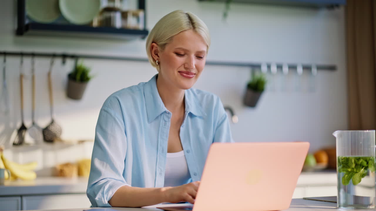 Tankefuld pige arbejder på computer og skriver idéer i køkkenet tæt på. Blondine der skriver