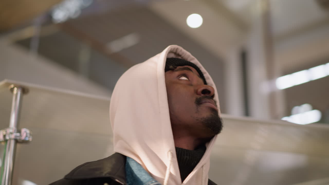 Close-up of young man with hoodie and beanie, looking upward with a curious expression, soft background lighting in modern shopping mall, slightly blurred people in the background