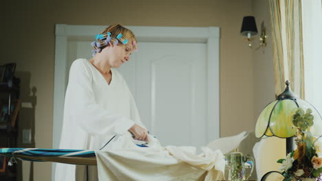 Woman-In-A-Bathrobe-With-Curlers-On-Her-Head-Ironing-Clothes