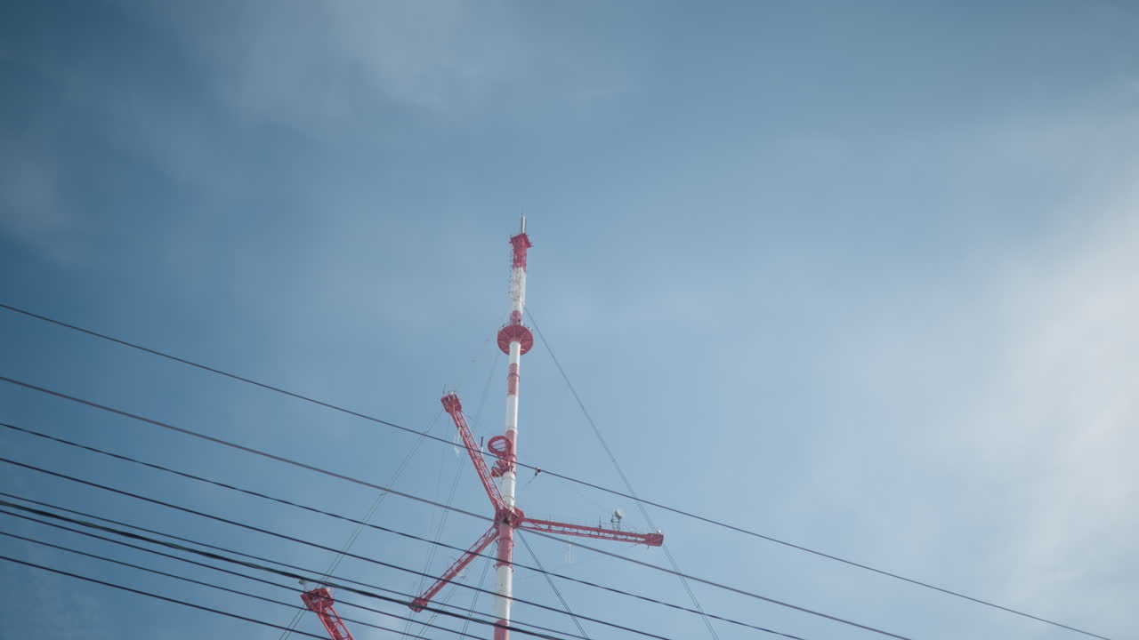 upward view across bus stop canopy toward red white transmission tower, power lines crisscross blue sky, urban winter transit scene framed by shelter roof, infrastructure focus for city commute