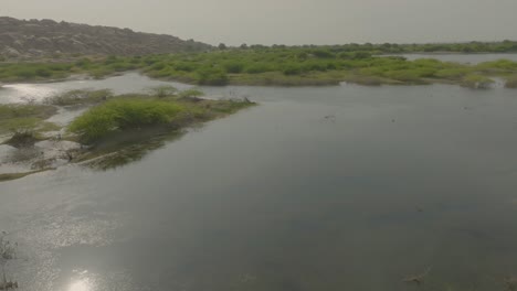 Un-Dron-Sobrevuela-Una-Presa-En-Nagarparkar,-Revelando-Sus-Tranquilas-Aguas-Y-Los-Contrastantes-Paisajes-áridos-De-Sindh,-Pakistán,-En-Una-Breve-Y-Serena-Instantánea