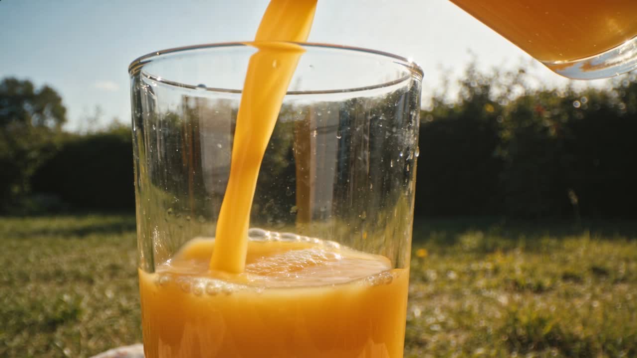 Kanne som tippes heller appelsinjuice i et gjennomsiktig glass på en plen i bakgården, fylles med skum