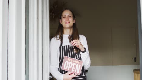 una camarera caucásica sonriente de pie en la puerta, sosteniendo un cartel abierto, mirando a la cámara