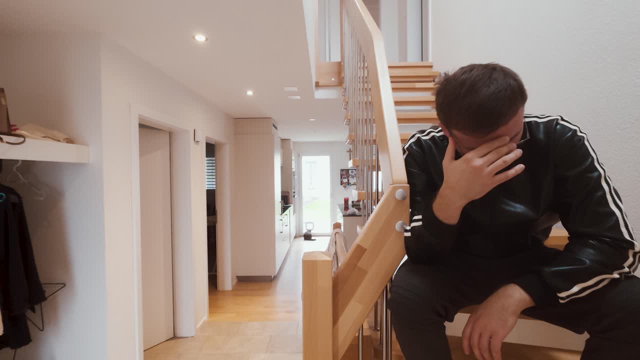 A man sits alone on the stairs in his apartment, covering his face in sadness and despair. Perfect for themes of loneliness, stress, depression, or emotional struggle in everyday life