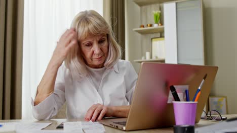 una anciana sentada en casa en una mesa calculada en una calculadora el costo por mes mira de cerca el gran número de cheques dudas y no entiende por qué tantos costos