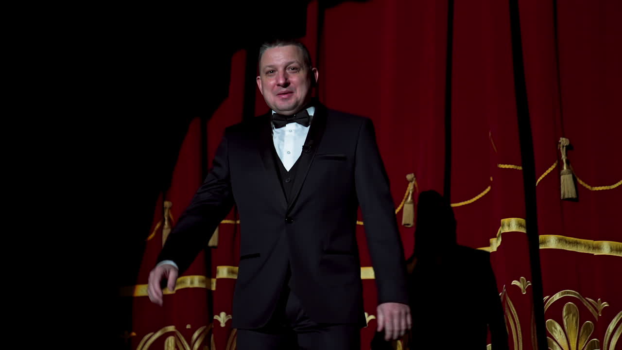Portrait of a host. Man actor in black costume appears on stage and begins the speech in bright light spot in the theater.