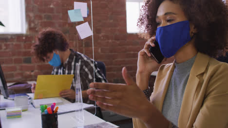 mujer de negocios de raza mixta hablando por teléfono sentada frente a la computadora con máscara facial