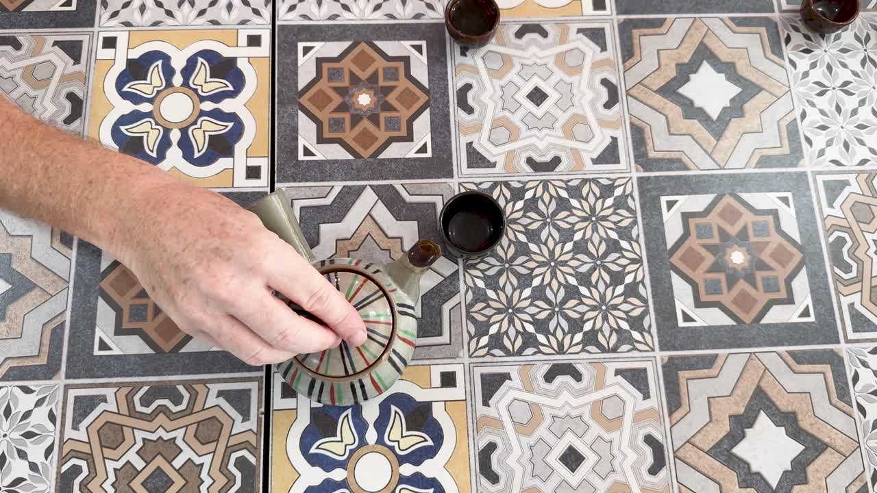 Hand opens ceramic teapot lid on colorful patterned tile table, overhead view, natural daylight