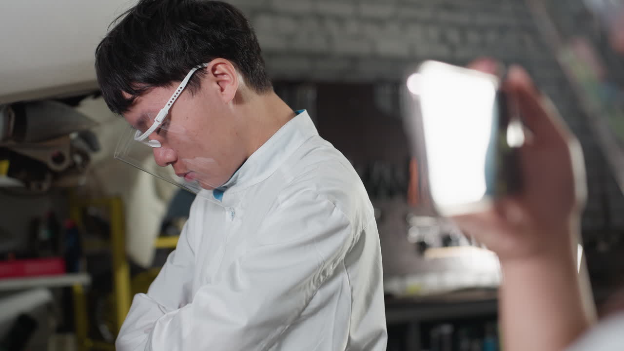 보호 안경을 입은 실험실 기술자가 손을 고 머리를 숙이고 자동차 작업장 배경에 있는 스마트폰의 클로즈업