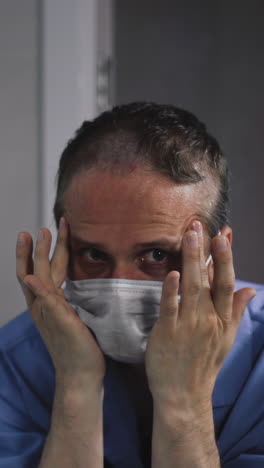 closeup of young uncut unshaven doctor, dressed in intensive care suit, in front of mirror, puts on protective mask, massages himself face, doing facebuilding, temples, gets scared, runs away