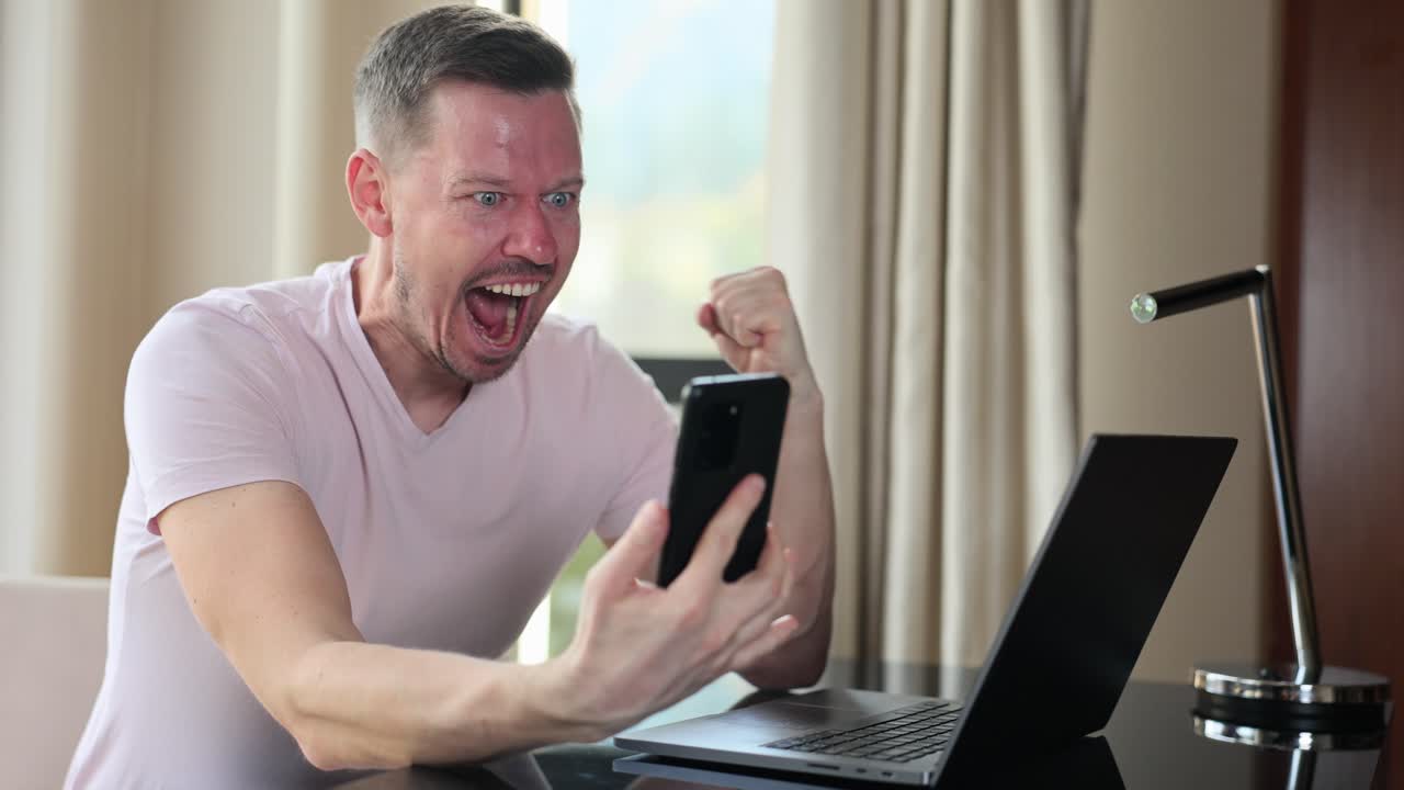 Excited man celebrating success with mobile phone and laptop