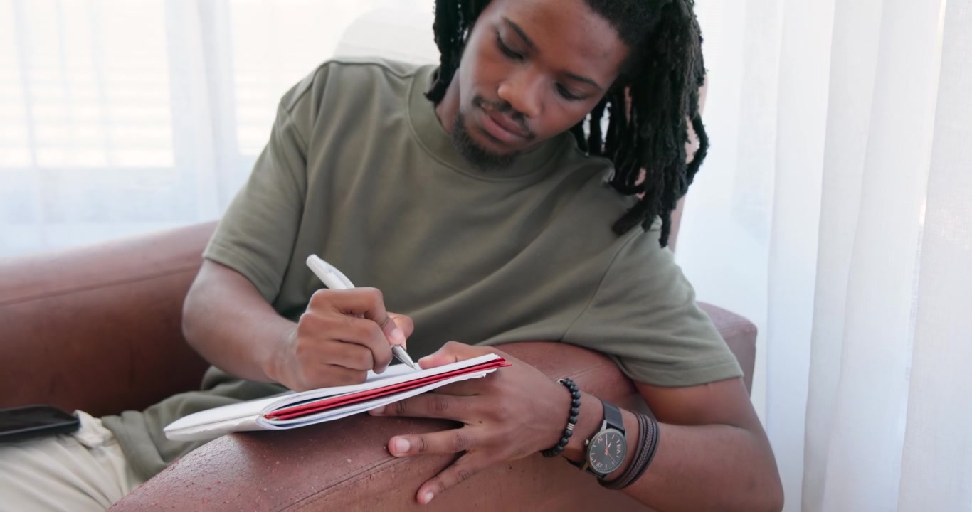 African American man in olive-tee checking phone, picking pen and writing in red notebook at home