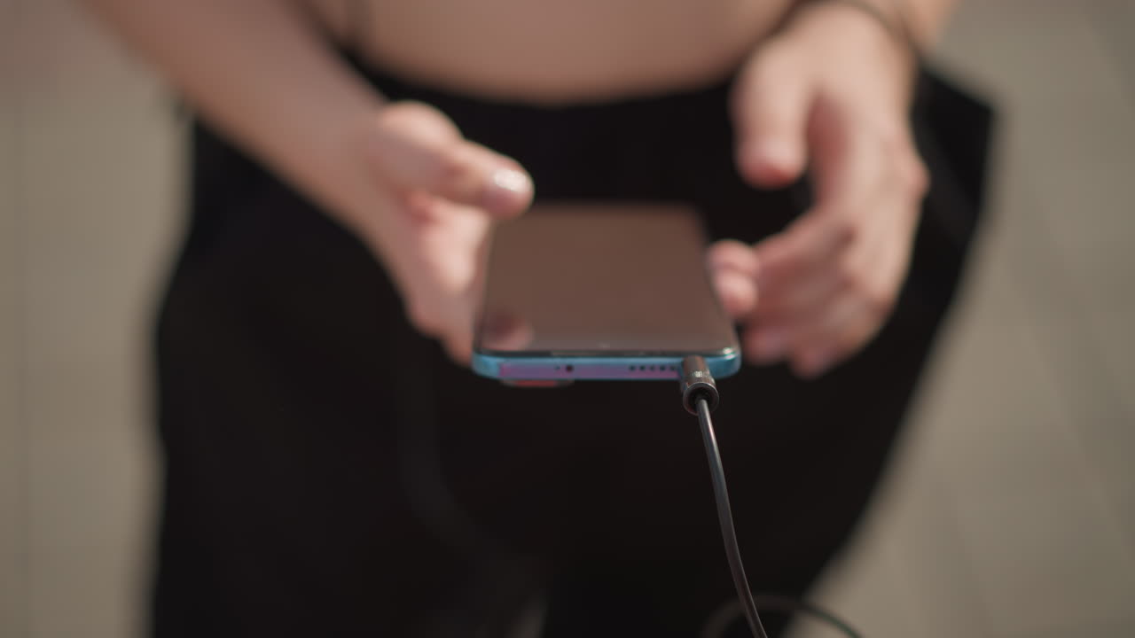 Giovane donna collega le cuffie al telefono all'aperto, torso e mani ritagliati, cavo delle cuffie inserito nel dispositivo, custodia protettiva del telefono, luce solare sul pavimento piastrellato, preparazione concentrata per la strada