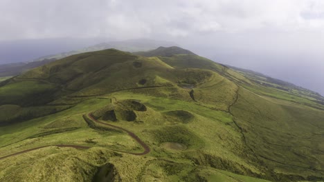 drone-dolly-view-of-vulcanic-mountains-covered-in-lush-green-vegetation-with-dirt-road,-cloudy-weather-in-São-Jorge-island,-the-Azores,-Portugal