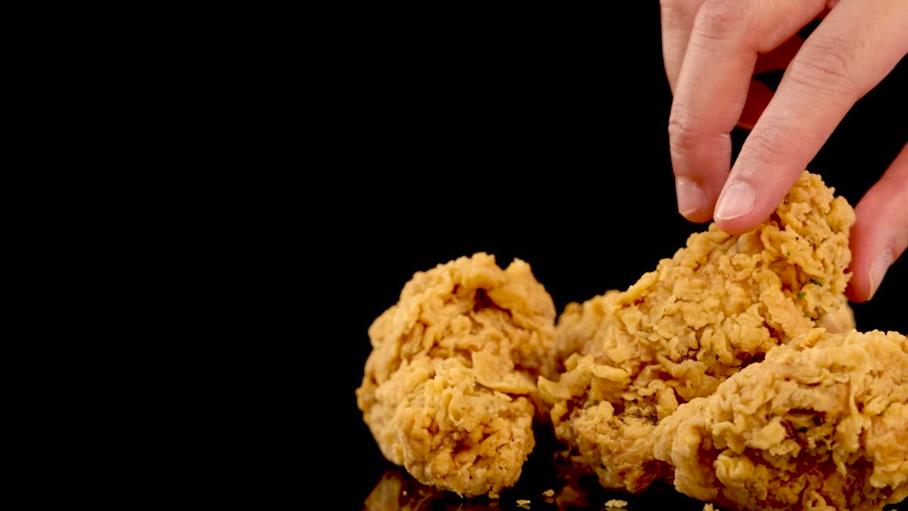 A hand selects a golden, crumbed fried chicken piece from a pile on a glossy black surface, with dramatic studio lighting and slow motion effect