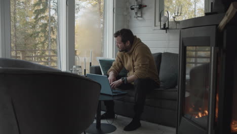 man with laptop works daytime job in front of fireplace in cozy small cabin home