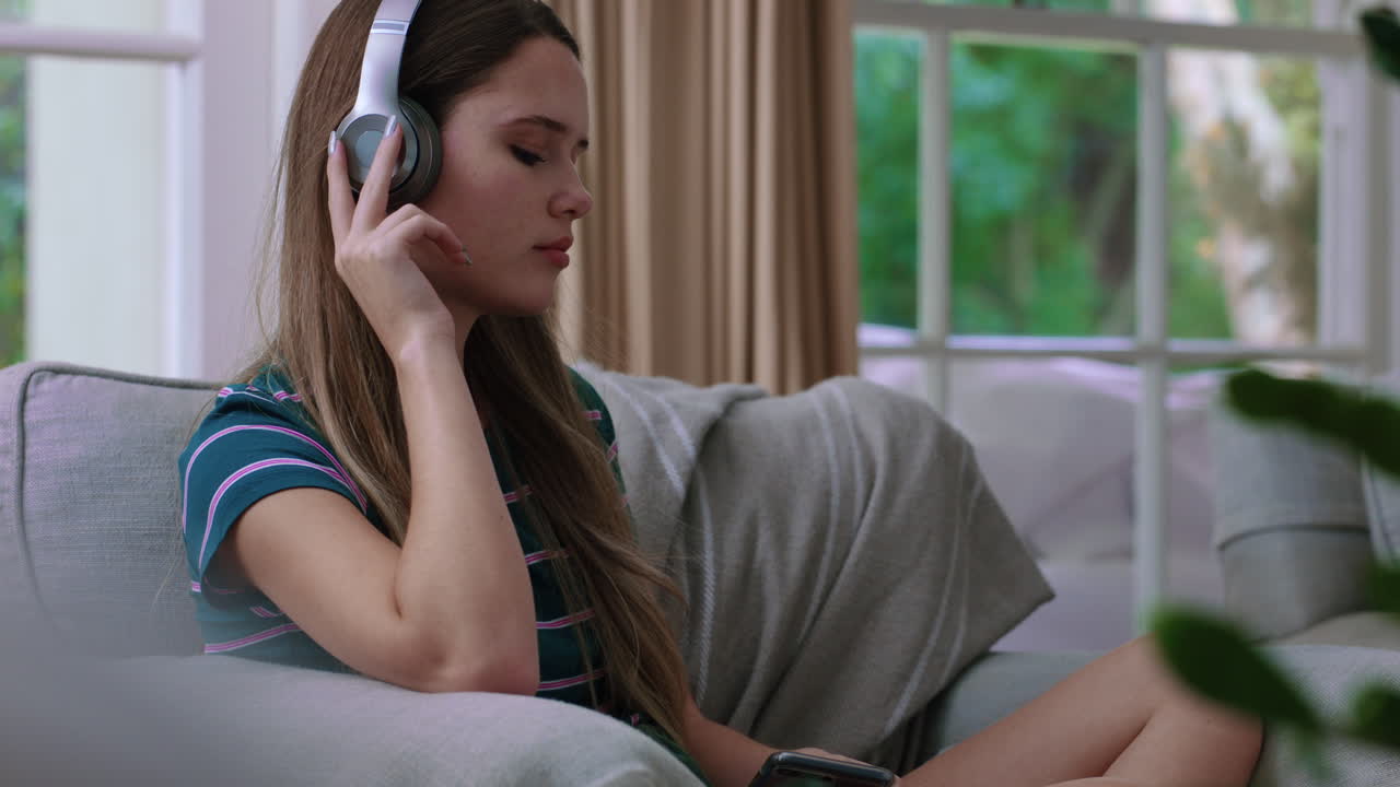 행복한 십대 소녀가 음악을 듣고 헤드폰을 착용하고 집의 소파에서 휴식을 취하고 스마트폰으로 문자 메시지를 보내고 소셜 미디어에서 라이프 스타일을 공유하는 것을 즐기고 있습니다.
