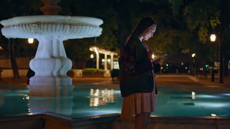 young woman scrolling mobile phone standing near city fountain late evening.