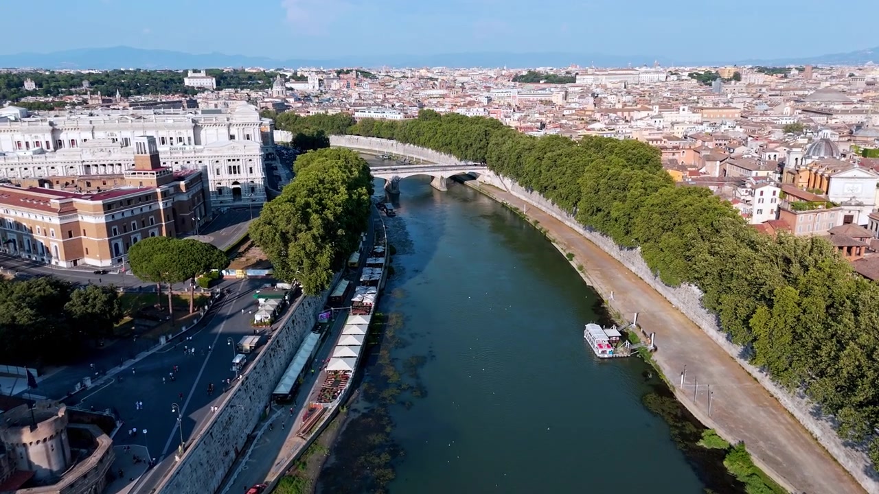 Castel Sant'Angelo costeggia il Tevere alberato mentre un sorvolo di drone diurno si muove verso Ponte Sant'Angelo e i tetti distanti della città.