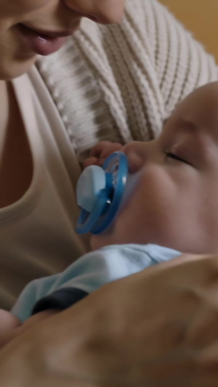 Vertical video: Cradling mother in cardigan after infant fussing, adjusting blue pacifier at home