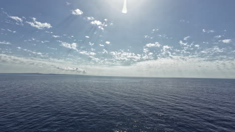 Speedy-drone-flight-over-the-sea,-looking-towards-the-sun-and-land-in-the-horizon-on-a-beautiful-day,-truck-shot-sidewise-on-a-cloudy-but-sunny-day-with-copy-space