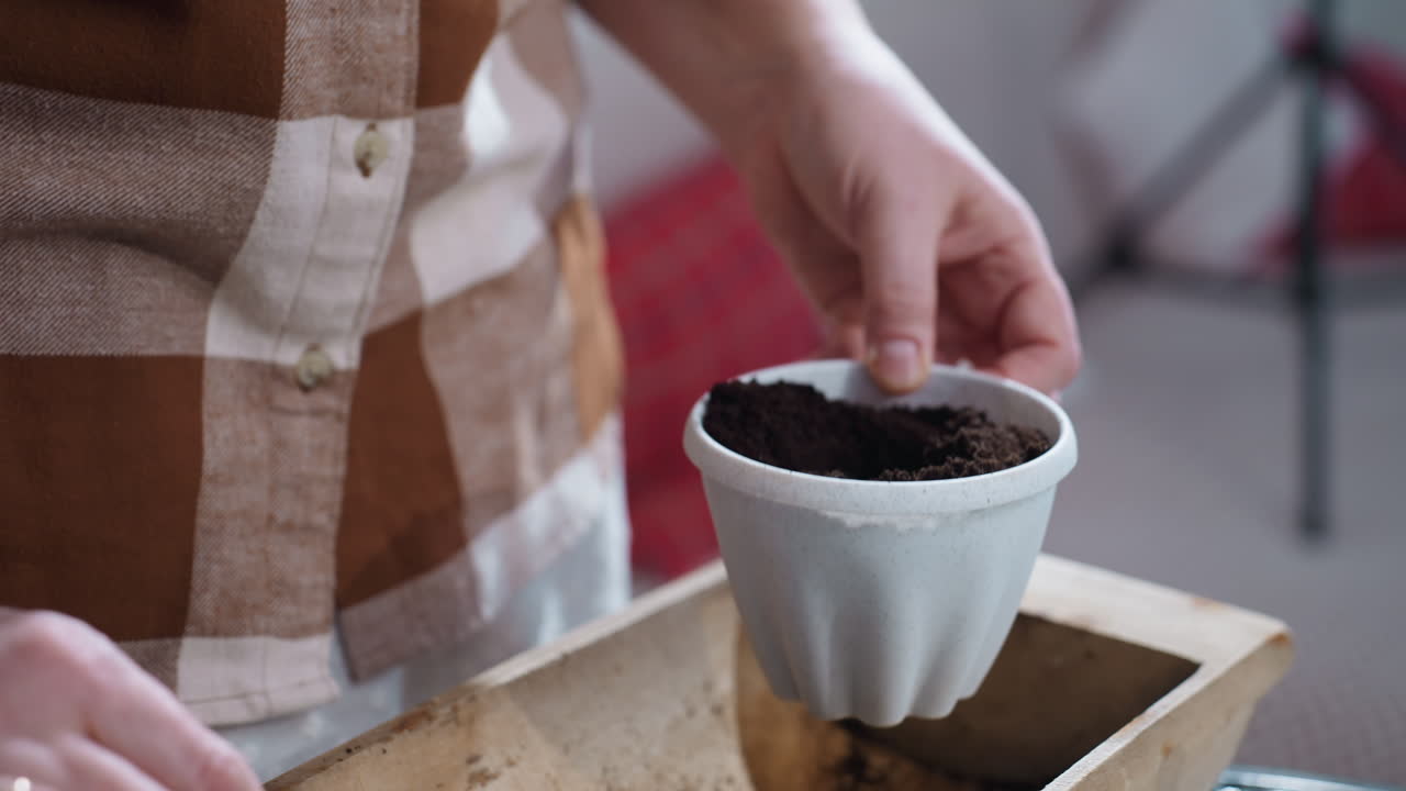 Lower view of gardener hands using small shovel to dig hole in plastic pot filled with dark soil and gently placing young plant with fresh roots into hole on indoor glass table with potted greenery