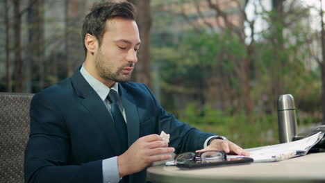 hombre de negocios relajado en traje revisando trabajos de papel o documentos en el portapapeles sentado en un parque de la ciudad durante el almuerzo comiendo bocadillos, volteando páginas y leyendo asintiendo
