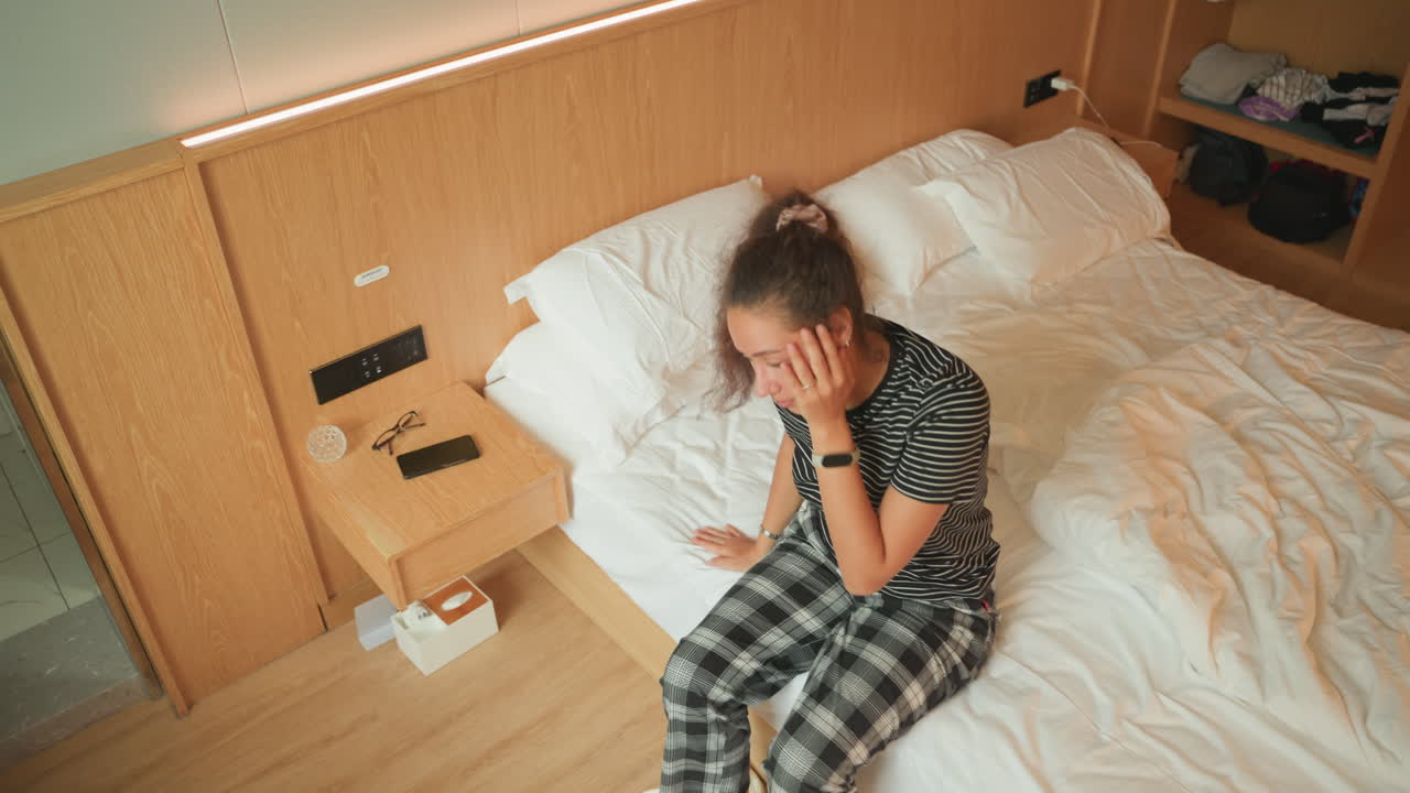 Una donna si sveglia nel letto d'hotel, si siede dondolando le gambe. Indossa vestiti a quadri, con una testiera in legno, un comodino con telefono e lampada, una morbida illuminazione a strisce LED, e un pavimento in legno. Allunga la mano verso la valigia e si alza.