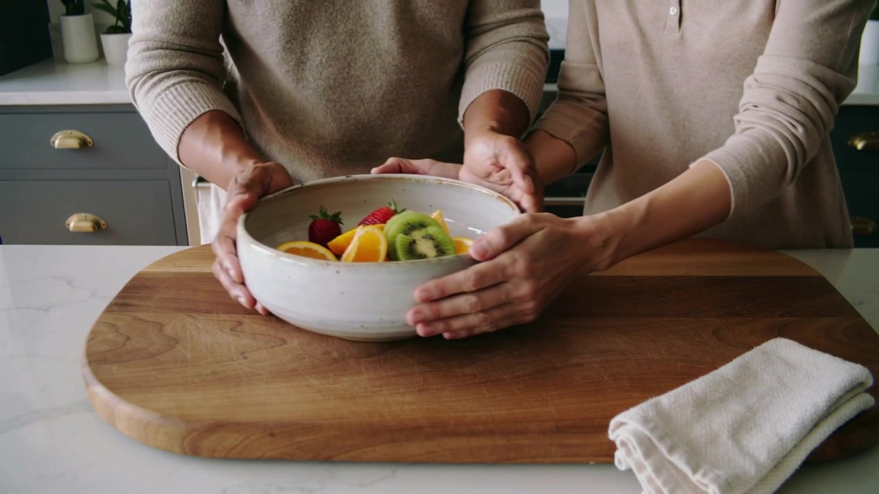 Lowering bowl as hands enter, two women wearing knit tops centering fruit on board in kitchen